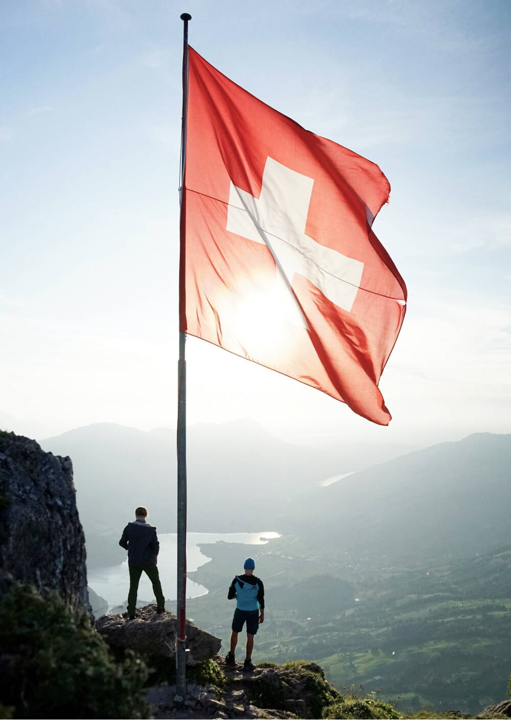 rectangle_273.jpg Ein Mann steht auf einer Bergspitze unter einer Schweizer Fahne und schaut über das beeindruckende Bergtal.